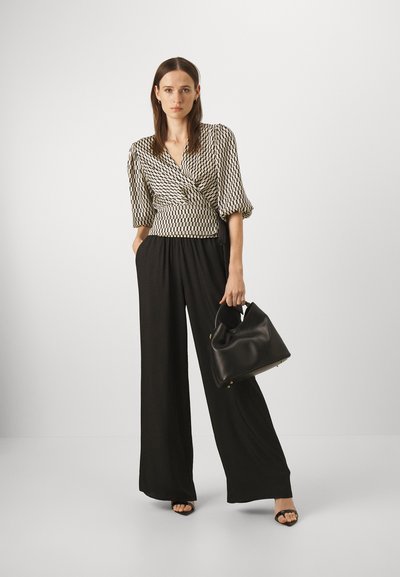 Svarte vide bukser kombinert med en mønstret omslagsbluse i krem og svart. Modellen bærer en svart lærveske med rundt design.