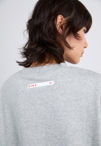 Sivi dukserica s istaknutom bijelom etiketom na kojoj piše "NIKE" crvenim slovima i malim Swoosh logom. Mekana tekstura, ležeran dizajn.