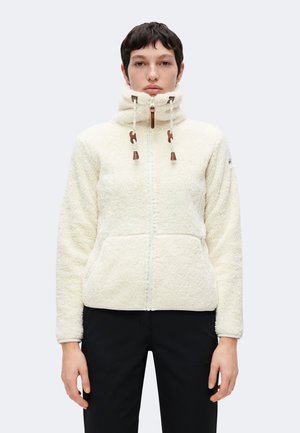 Persona con una chaqueta de forro polar blanca con cremallera, cuello alto y cordones, combinada con pantalones negros, de pie frente a un fondo claro.
