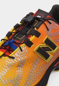 Chaussure de sport orange vif avec logo noir et jaune, lacets élastiques noirs avec verrou, languette bleue et motifs texturés réfléchissants.