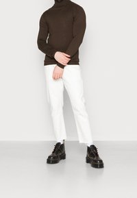 Maglione a collo alto marrone, pantaloni bianchi a gamba dritta e stivali neri con lacci e cuciture gialle. Tessuto liscio e design aderente.