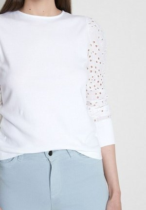 Femme portant un haut blanc avec des manches transparentes à pois et un pantalon bleu clair sur un fond uni.