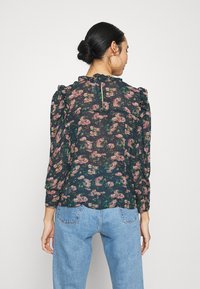 Blusa a fiori verde scuro con dettagli rosa, spalle arricciate e apertura a chiave sul retro. Abbinata a jeans a vita alta blu chiaro.