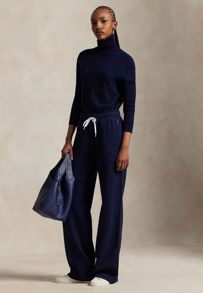 Vrouw met gevlochten haar, gekleed in een marineblauwe coltrui en broek met wijde pijpen, die een grote marineblauwe handtas vasthoudt, staand tegen een neutrale achtergrond.