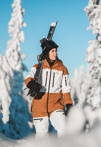 Ski jacket in brown and white color block design with black accents. Equipped with zippers and pockets, worn by a person holding skis.