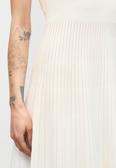 Αριστερό χέρι με λεπτομερείς τατουάζ δίπλα σε λευκή ριμπ πλεκτή φούστα με ψηλή μέση, πάνω σε απλό ανοιχτόχρωμο φόντο.