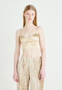 Satin cropped top u svijetlo zlatnoj boji, s tankim trakama i nabranim dizajnom na grudima. Uparen s bež prugastim plisiranim hlačama.
