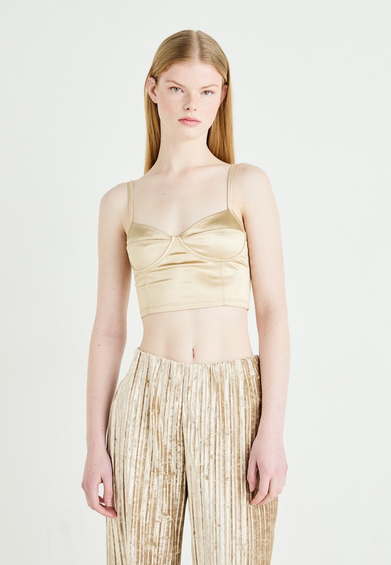 Satin cropped top u svijetlo zlatnoj boji, s tankim trakama i nabranim dizajnom na grudima. Uparen s bež prugastim plisiranim hlačama.