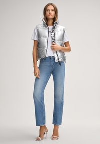 Srebrna puchowa kamizelka z wysokim kołnierzem, na zamek błyskawiczny, z czarnymi akcentami, noszona na białej koszulce, w zestawieniu z jasnoniebieskimi jeansami prostymi i srebrnymi szpilkami.