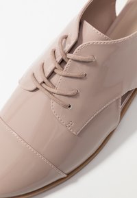 Chaussure en cuir verni rose clair brillant avec laçage frontal, bout arrondi et design minimaliste. Présente des coutures discrètes et une texture lisse.