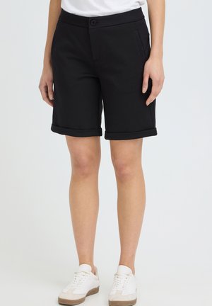 Person trägt schwarze umgekrempelte knielange Shorts mit Taschen, weiße Turnschuhe und ein hineingestecktes weißes Hemd vor einfarbigem Hintergrund.