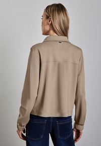 Veste cropped beige en tissu doux, dotée d'un col, de poignets élastiques et d'une couture horizontale dans le dos. Un pantalon en jean bleu est visible.