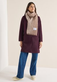 Viininpunainen takki taskuilla, yhdistetty tekstuuriin beigellä huivilla, jossa on hapsut. Farkkuleveälahkeiset housut ja vaaleat kengät täydentävät asun.