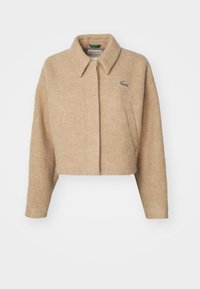 Manteau en laine beige coupé avec un col, deux poches avant et un accent logo Lacoste sur le côté supérieur gauche. Texture douce.