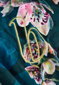 Kvetinová látka s ružovými a zelenými orchideami na tmavo-modrom pozadí, s vyšitým zlatým vláknom vytvárajúcim kurzívne písmeno "b".