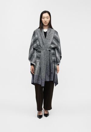 Woman wearing a black and white patterned belted coat over black pants and black pointed heels, standing against a plain white background.