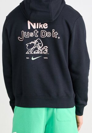 Osoba ubrana w czarną bluzę z kapturem Nike z napisem „Just Do It”, rysunkiem Snoopy’ego i zielonym logo Nike, połączone z jaskrawozielonymi szortami.