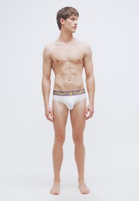 Λευκά εσώρουχα slip με ελαστική ζώνη μέσης σε μοτίβο μωβ και χρυσού. Το μοντέλο στέκεται με τα χέρια στα πλάγια, χωρίς παπούτσια, και κοιτά ζωντανά μπροστά.