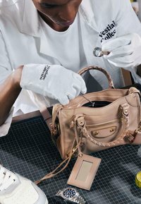 Personne portant des gants blancs et un manteau blanc utilise une loupe pour inspecter un sac à main en cuir beige sur une surface quadrillée.