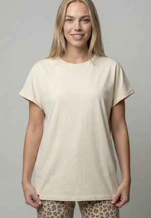 Blonde Frau, die lächelt, trägt ein schlichtes beiges T-Shirt mit kurzen Ärmeln und Leopardenmuster-Hose, steht vor einem neutralen grauen Hintergrund.