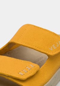 Keltainen mokkanahkainen sandaali kahdella säädettävällä tarranauhalla, kontrastiväriset ompeleet ja teksturoitu kumipohja. Brändinimi 'VEJA' kaiverrettuna sivulle.