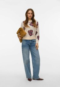 Krēmkrāsas džemperis ar ziedu rakstu, rozā toņos, kombinēts ar augsta vidukļa zilām džinsa biksēm un brūnām apaviem ar smailiem pirkstiem. Rokās tur brūnu rokassomiņu.