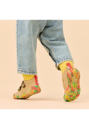 Kind trägt blaue Jeans, gelbe Socken und beige Schuhe mit bunten, strukturierten Sohlen und macht einen Schritt nach vorne auf einem hellbeigen Hintergrund.