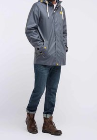 Man som bär en mörkgrå regnrock med gula knappar, uppkavlade mörkblå jeans och bruna snörstövlar, står med händerna i fickorna.