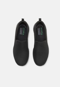 Zapatos negros sin cordones con una parte superior texturizada perforada, suela plana y dos tiradores para facilitar su puesta. El interior presenta una marca impresa en verde.