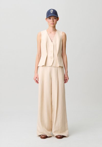 Gilet in lino beige con scollo a V e due bottoni, abbinato a pantaloni beige a gamba larga con sottili righe verticali e scarpe marroni.
