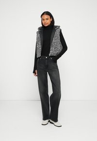 Černý top s dlouhým rukávem kombinovaný se šedou vestou s bílým abstraktním vzorem, stylizovaný s černými džíny s vysokým pasem a bílými botami.