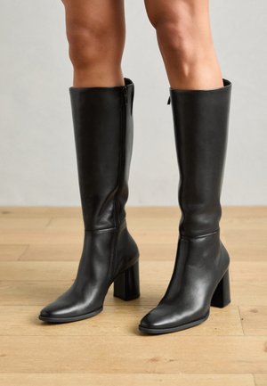 Schwarze Leder-Stiefel bis zum Knie mit quadrater Zehenform, hohem Blockabsatz und seitlichem Reißverschluss, vor einem Hintergrund aus Holzboden.