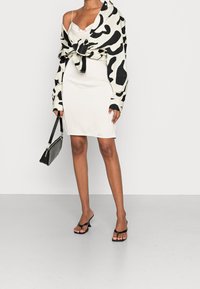 Bílá těsně přiléhající šaty kombinované s volnou, uzlou černobílou košilí se vzorem, černou kabelkou a černými šlapkami.