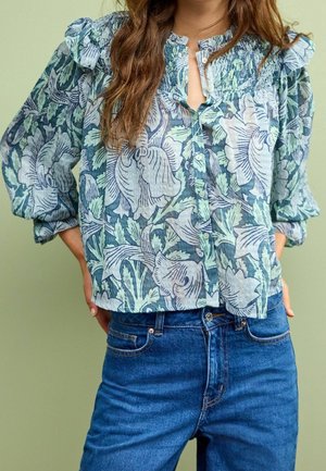 Vrouw die een blauwe blouse met bloemenpatroon draagt, met ruches op de schouders en lange pofmouwen, gecombineerd met een high-waisted blauwe jeans tegen een groene achtergrond.