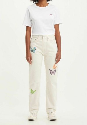 Personne portant un jean blanc avec des imprimés papillons colorés et un t-shirt blanc uni à manches courtes, debout devant un fond clair.