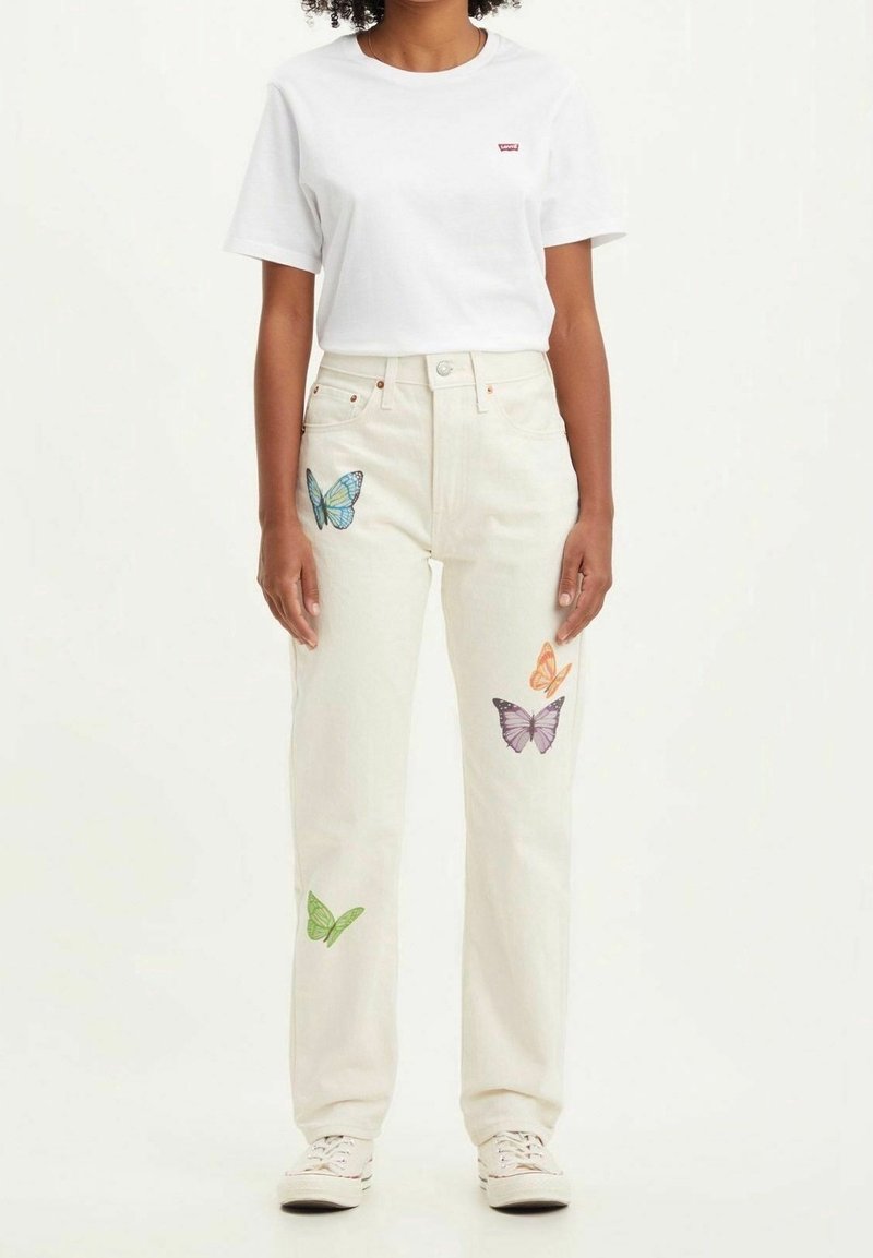 Personne portant un jean blanc avec des imprimés papillons colorés et un t-shirt blanc uni à manches courtes, debout devant un fond clair.