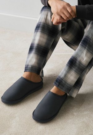 Persoană așezată cu mâinile încleștate, purtând pantaloni carouri negri și gri și papuci gri închis, fără șiret, pe un covor deschis la culoare.