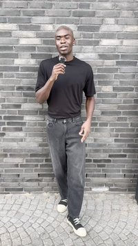 T-shirt preta de manga curta, calças baggy cinzentas e sapatilhas pretas com solas brancas. Pessoa a segurar um objeto escuro, de pé contra uma parede de tijolos cinzentos.