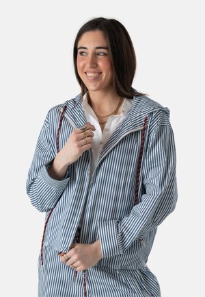 Joven sonriente, vestida con chaqueta con cremallera y pantalones a juego de rayas verticales en azul y blanco, con varios collares dorados superpuestos y anillos.