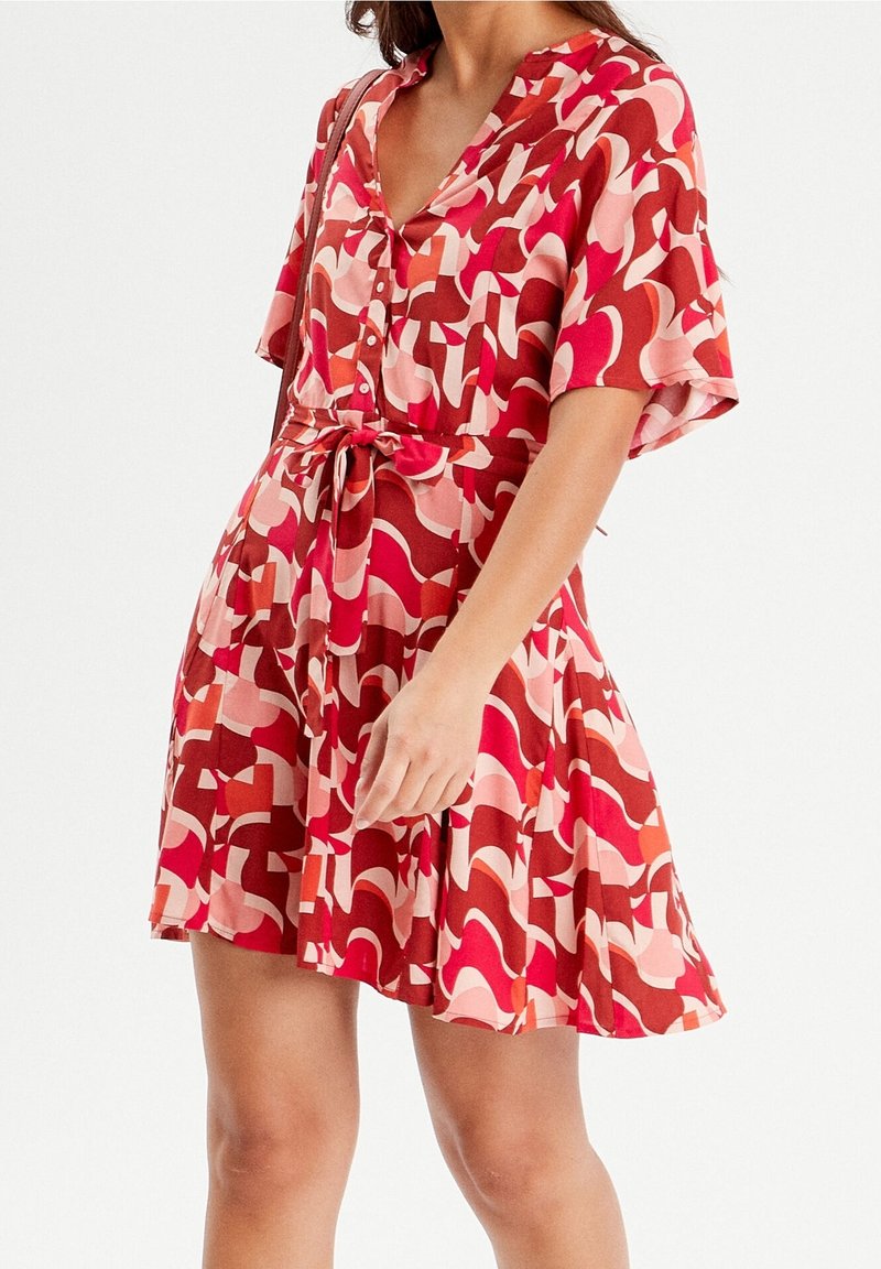 Femme portant une robe à manches courtes rouge, rose et blanche à motifs, avec des boutons et une ceinture nouée à la taille.
