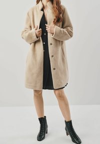 Cappotto in misto lana beige con colletto alto, chiusura a cinque bottoni e due tasche laterali. Abbinato a un vestito nero plissettato e stivaletti neri.