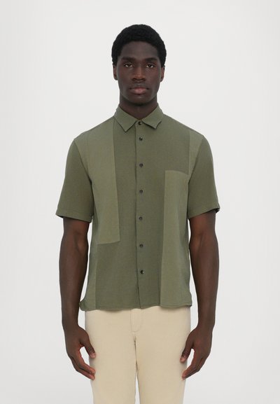 Short-sleeved, olive green button-up shirt with a textured fabric, featuring a patch pocket and contrasting panels in different shades.