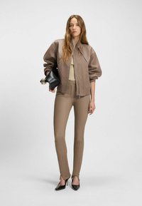 Ruskea pommitakki, jossa on pullistuvat hihat ja nahkaaksentit, beigen sävyinen toppi, istuvat beigen väriset housut vetoketjuyksityiskohdilla ja mustat korkokengät.