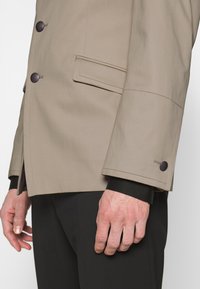 Beige jacka med skräddarsydd passform, som har svarta knappar, en bröstficka och en jämn textur. Ytterligare svarta detaljer vid ärmsluten.