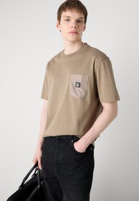 Cămașă t-shirt din bumbac bej, cu un buzunar într-o nuanță de bej deschis, având un patch cu logo negru. Asortată cu blugi negri și o geantă neagră.