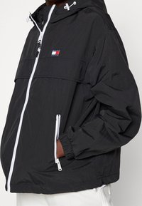 Zwarte windbreaker jas met witte accenten, ritszakken en elastische manchetten. Het heeft een logodetail op de borst en een verstelbare capuchon.