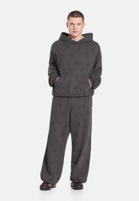 Ciemnoszara bluza z kapturem i pasujące szerokie spodnie z miękkiego materiału z delikatnym wzorem. Posiadają przednią kieszeń i kaptur.