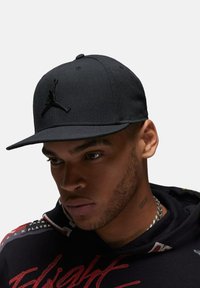 Sort baseball cap med flad skygge og broderet Jordan-logo. Materialet er solidt og tekstureret, med en struktureret form.