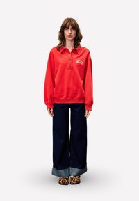 Sweatshirt rouge à col et boutons, associé à un jean en denim foncé à jambe large avec un ourlet roulé. Des chaussures imprimé léopard complètent la tenue.