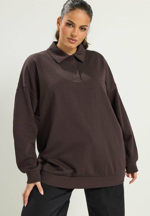 Une femme porte un sweat-shirt oversize marron foncé à col et à patte de boutonnage, ainsi qu’un pantalon noir, sur un fond de studio clair.